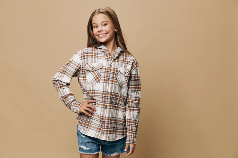 Girl smiling and posing with hand on hip wearing plaid shirt and denim shorts standing against beige background. Casual clothing and confident posture with youth style.