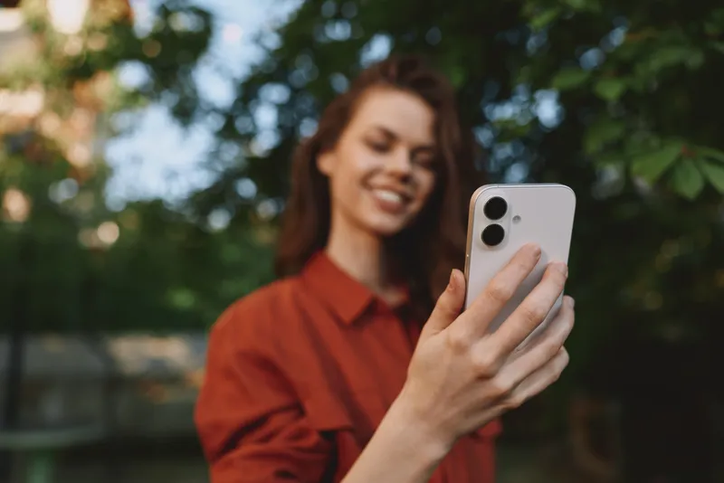 Smiling woman taking a selfie outdoors with a smartphone, wearing a red shirt. Natural daylight and green background convey lifestyle, wellness, and everyday confidence in a bright, relaxed moment