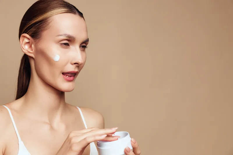Young woman with long blonde hair applies moisturizer cream on her face while holding a white jar against a plain beige background in a skincare routine scene.