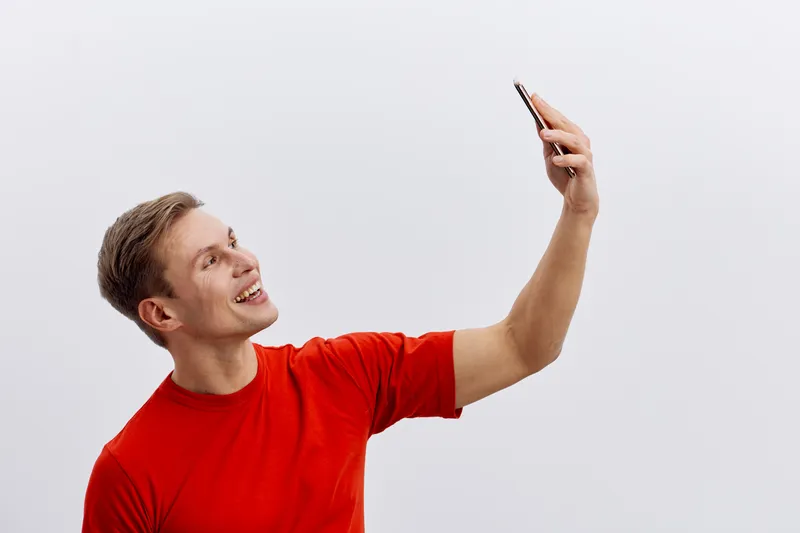 Smiling young man with short hair wearing casual red T-shirt taking a selfie with smartphone, looking happy and cheerful, isolated on plain white background, studio portrait. People lifestyle concept