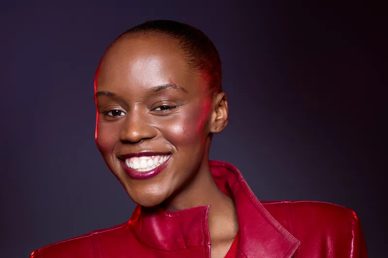 Confident smiling black woman with dark skin and short hair wears red leather jacket against purple background. Studio portrait of young African female model with bright makeup and positive mood.