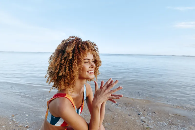 Smiling woman in a striped swimsuit enjoys a sunny day at the seaside, standing on wet sand near calm ocean, capturing carefree summer mood and coastal relaxation