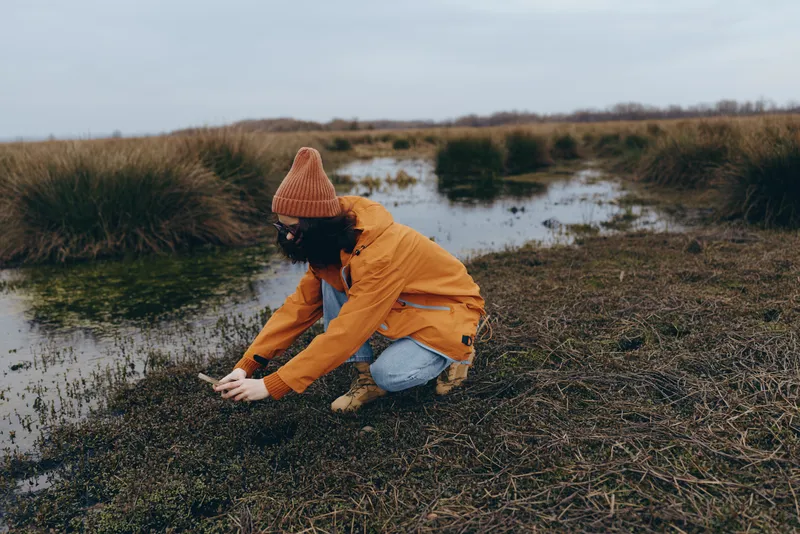 Woman in outdoor nature lifestyle, kneeling by a riverbank, wearing an orange jacket and knit hat, touching wet grass, soft daylight over calm marshland, candid lifestyle moment