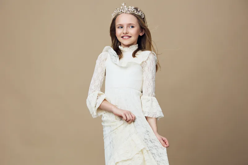 Elegant young girl in a white lace dress with a crown, smiling joyfully against a soft beige background, perfect for fashion and beauty concepts