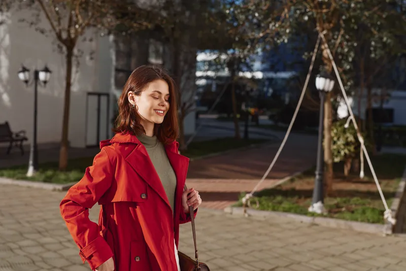 A young woman in a bright red coat walks outdoors, smiling softly as she carries a small bag along a sunlit plaza.
