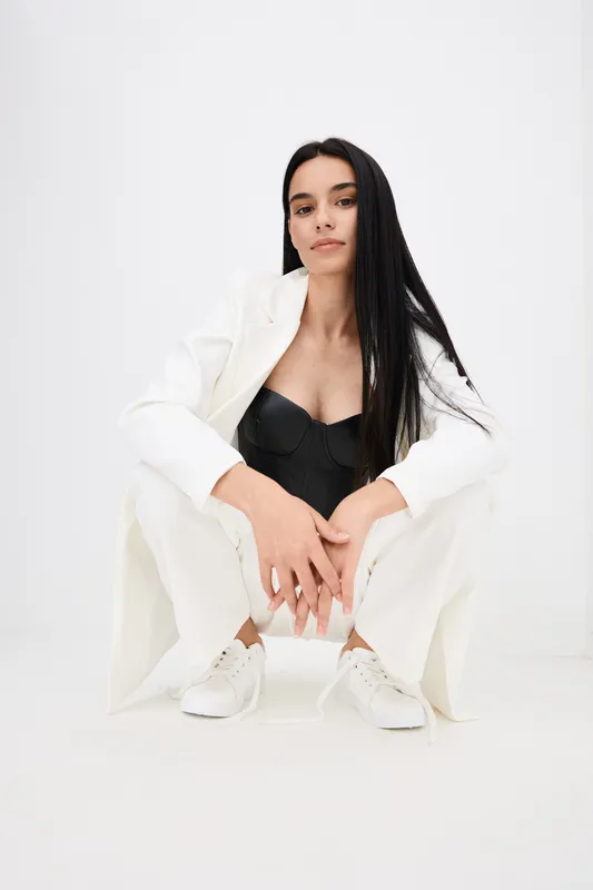 young woman wearing white oversized jacket and pants with black bustier, sitting in stylish pose against white background, casual modern fashion look.