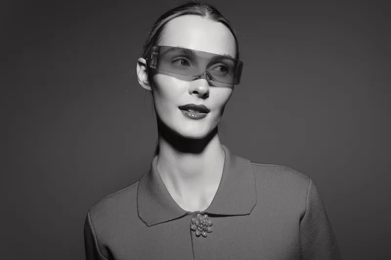 Slim Caucasian woman with fair skin wears futuristic translucent visor and textured top, posing confidently against dark studio background. Concept of fashion and innovation.