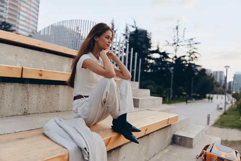 Stylish woman in business attire sits on urban steps, reflecting on her career in a modern cityscape with buildings and greenery in the background.