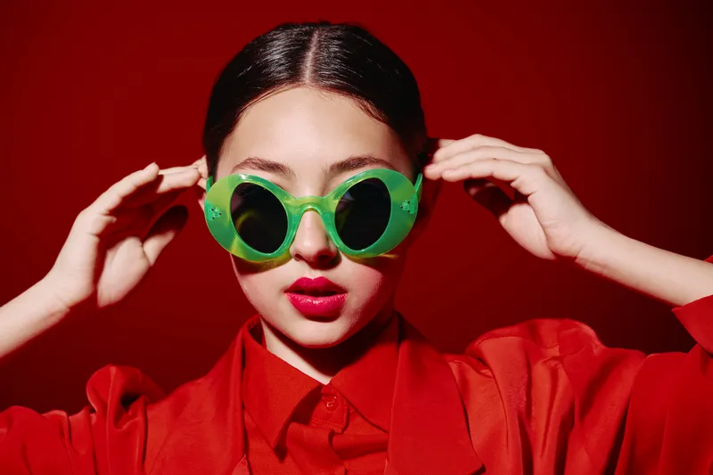 Young woman in bright red outerwear wears oversized green sunglasses. The glossy background complements the fat bold lips makeup. Fashion and beauty creative concept shot.