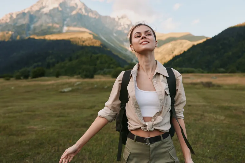 Young woman outdoors in a scenic mountain valley, wearing a tied beige shirt over a white top and a backpack, arms open, feeling confident and free today.