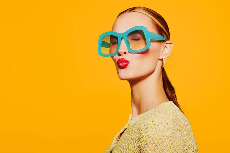 Stylish woman with slicked hair and oversized blue glasses blows a kiss on bright yellow background. Concept of fashion, beauty, playful attitude, and vibrant color contrast.