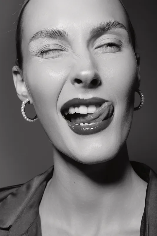 Caucasian woman with fair skin and minimal makeup poses in black and white close-up portrait, winking and licking lips. Concept of beauty, expression, and monochrome fashion photography.