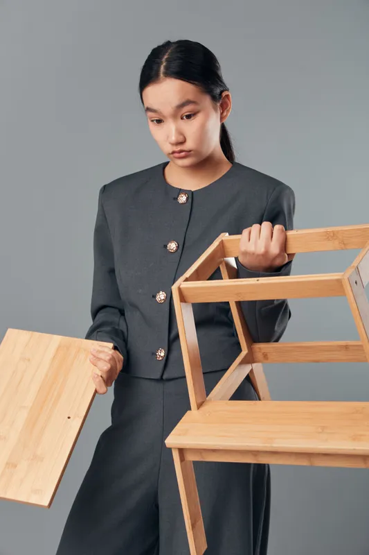 Young woman holding wooden chair and plank with thoughtful expression in studio, casual gray outfit and minimal background, furniture assembly or repair concept with focus on wood material.