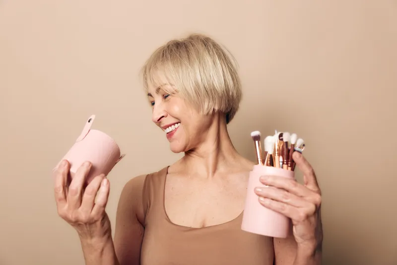 Smiling mature woman with short blonde hair holds pink containers with makeup brushes and cosmetics. Beige background highlights beauty routine concept, self-care, and joyful lifestyle.