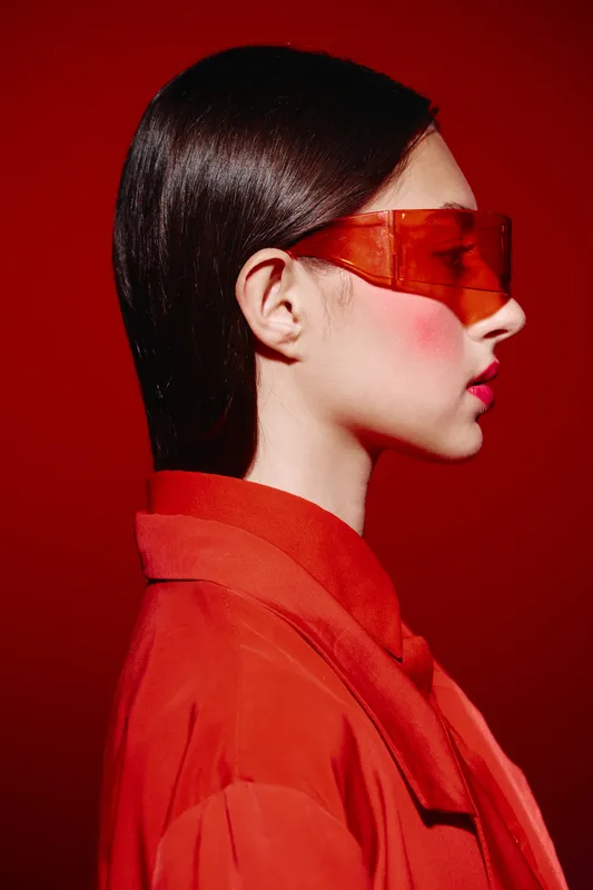 Profile of a stylish woman in vibrant red oversized jacket and orange sunglasses against matching red background. Concept for bold seasonal fashion and beauty creativity.
