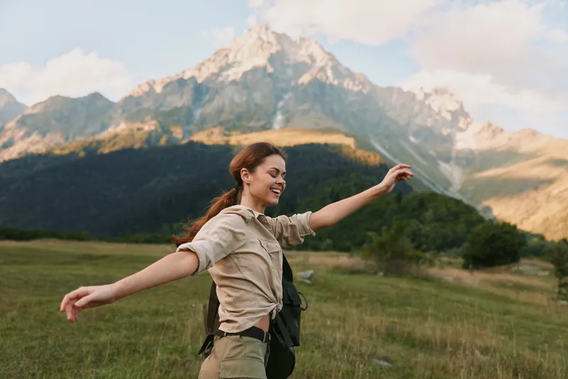 Young woman with long hair stands in a grassy valley among mountains, arms wide open and a bright, carefree smile, conveying freedom and outdoor adventure.