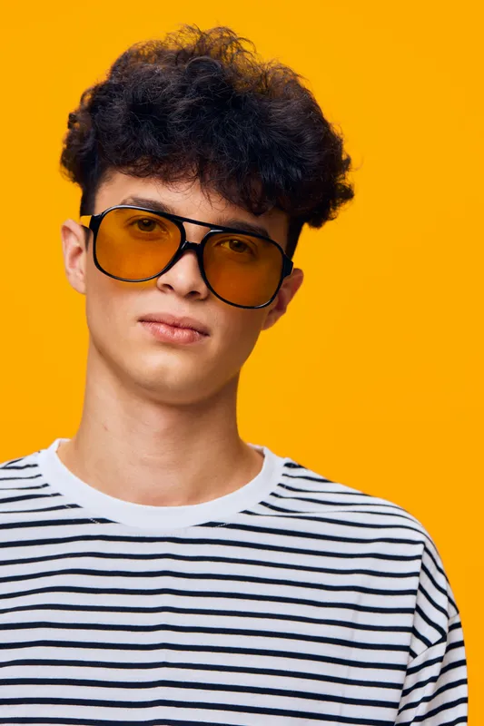 young man wearing oversized yellow sunglasses and striped shirt posing confidently against a vibrant orange background with curly hair style