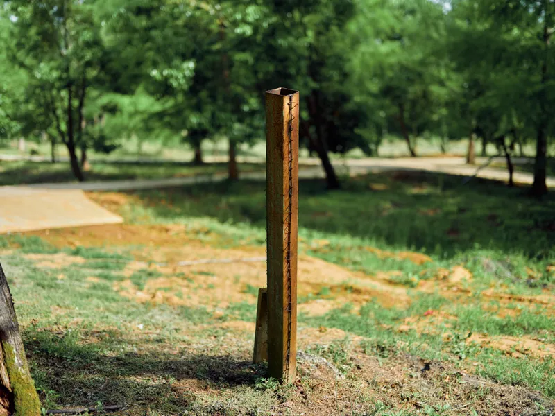 Post metal rusty park grass trees nature pole outdoors with weathered iron stake standing in sunlit grassy area, shallow depth of field and blurred woodland background.