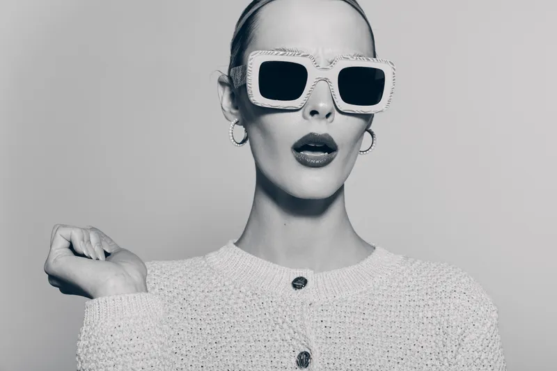 Stylish white woman with pale skin wearing large sunglasses and earrings poses with raised hand in monochrome studio photo. Fashion concept and confident attitude.