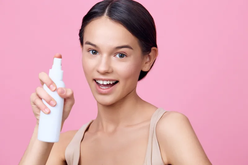 Young woman promoting skincare product, smiling and holding white spray bottle against pink background, demonstrating healthy skincare routine