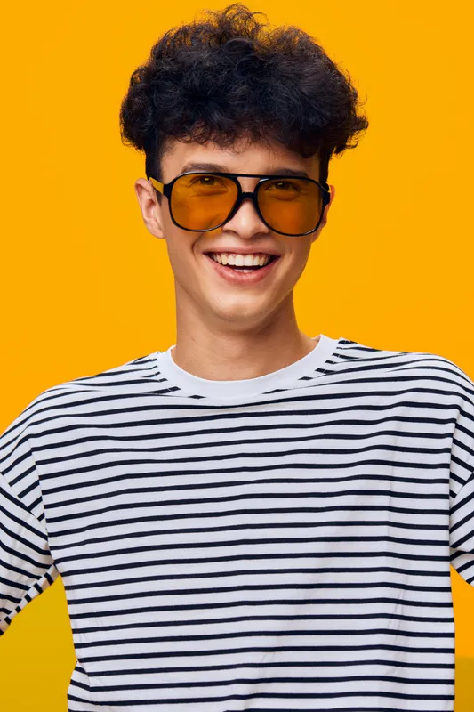 smiling man wearing yellow sunglasses and striped shirt with curly hair on a vibrant orange background, expressing happiness and youthfulness in casual style