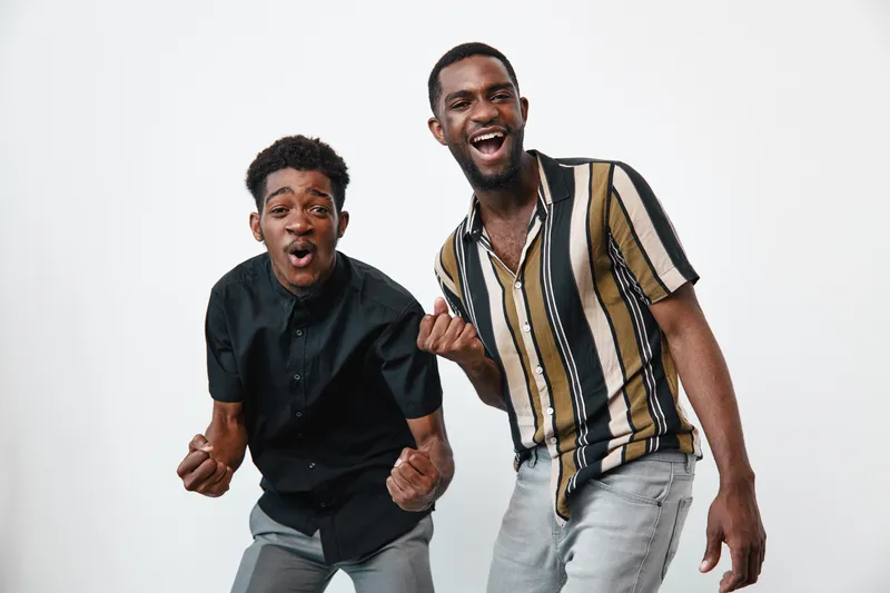 Two young black men with dark skin expressing excitement and joy in casual clothing against white background. Friends celebrating, smiling, and showing energetic emotions together.