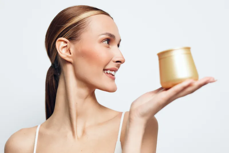 Smiling woman in profile holds a gold cream jar, displaying a skincare product against a clean white background, emphasizing minimal, premium beauty with calm aesthetic for skincare.
