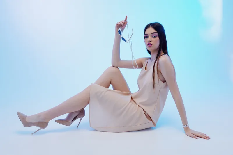 Young woman in a beige dress and high heels poses seated on the floor against a light blue background. Concept of beauty, fashion, elegance, and modern style.