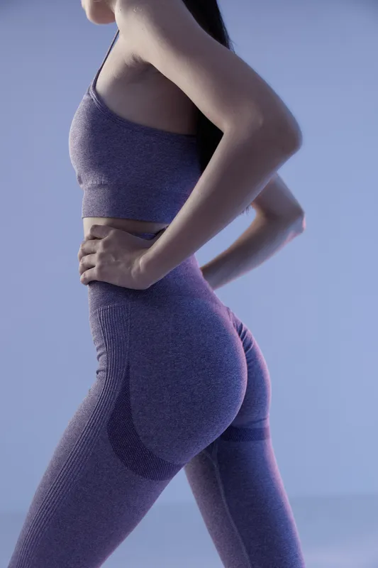athletic woman sport fitness wearing seamless purple activewear, posing with hands on hips in minimalistic studio with soft blue lighting and shadow play.