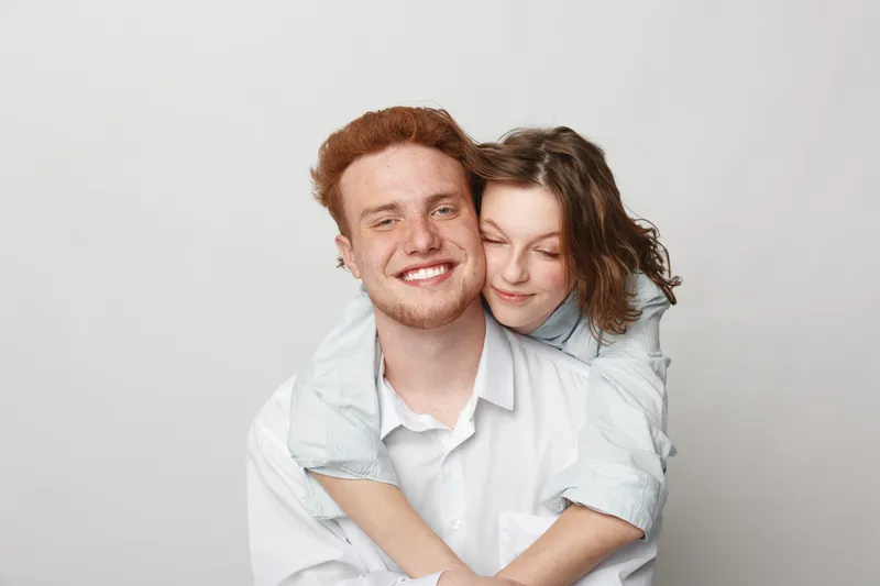 Happy young couple embraces and smiles with closed eyes in casual white shirts on light background. Concept of love, affection, friendship, and positive emotions between people.