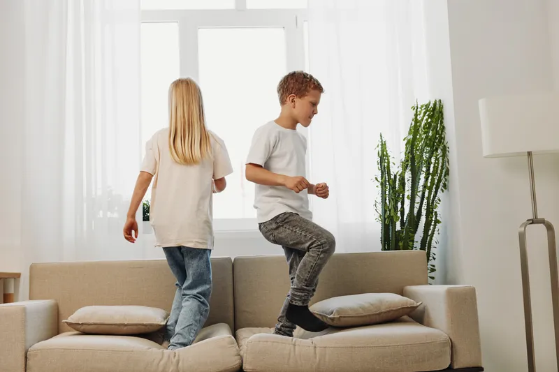 Two children wearing casual clothes jump and play on a beige sofa in a bright living room with large windows and modern decor. Concept of childhood fun and energy.