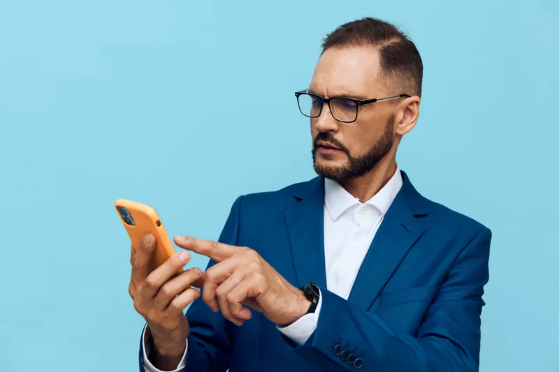 Professional man in a blue suit on a solid background, focused on his smartphone, exuding confidence, curiosity, and composure in a modern business setting.