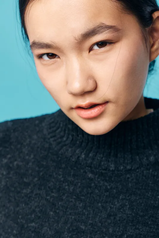 Young woman portrait with natural makeup and black sweater on blue background. Female model looking at camera with neutral expression, closeup of face and skin texture in studio.