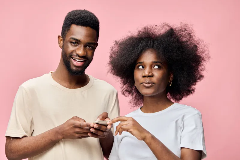 Young Black couple with stylish hairstyles, smiling and using smartphone against a pink background, representing modern relationships and technology