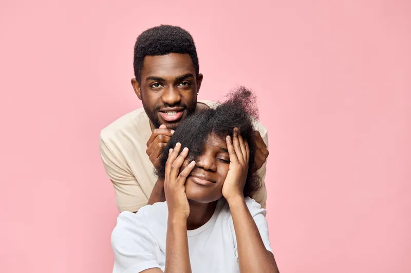 Young Black couple sharing a joyful moment, with the man gently holding the woman s hair, set against a soft pink background, capturing their playful and loving expressions