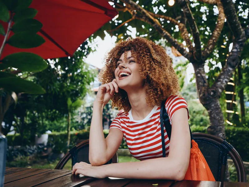 Joyful young woman with curly hair sits at an outdoor cafe, smiling warmly as sunlight glints through leaves and a red umbrella frames a vibrant, candid moment.