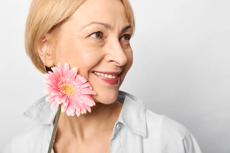 Smiling older woman portrait holding pink gerbera flower near face on white background gentle beauty concept fresh spring mood soft light minimalist studio closeup at eye level calm