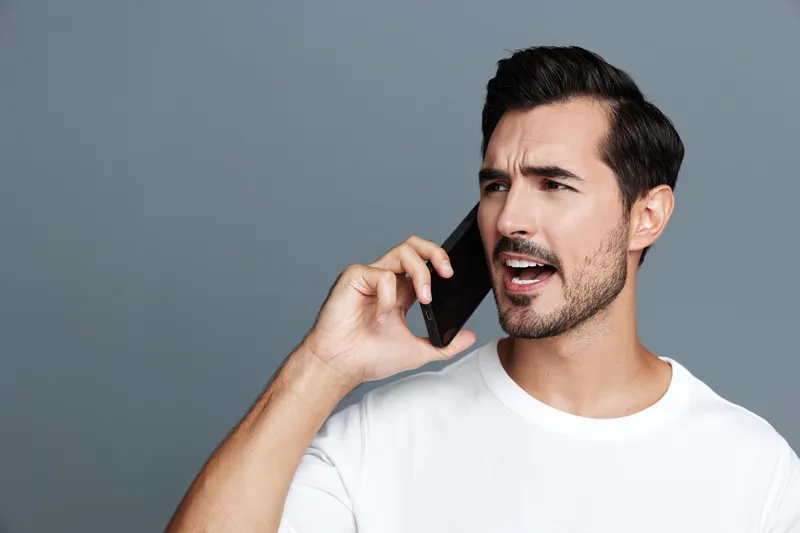 Young man in a white t-shirt talking on a mobile phone, expressing frustration or impatience against a plain gray background. Lifestyle communication concept.