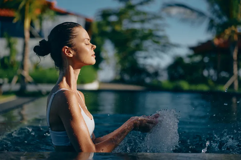 A young woman with a brunette bun stands at the pool edge, eyes closed as water splashes, conveying calm focus and carefree summer relaxation.
