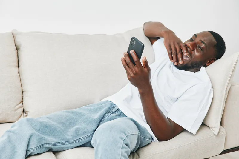 A relaxed young African American man in casual white t shirt and denim jeans laughing while using a smartphone on a cozy couch at home