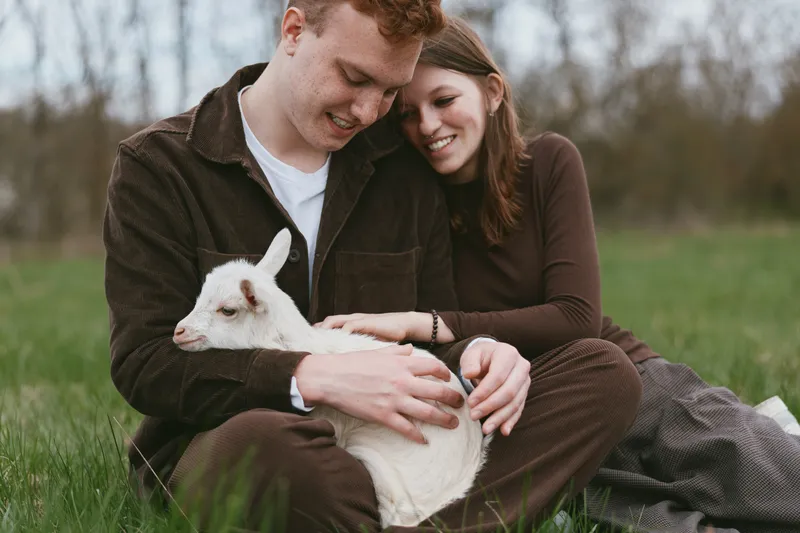 Two smiling individuals cuddle a white rabbit in a grassy field, sharing a calm, intimate moment outdoors. A wholesome lifestyle concept highlighting care, companionship, and connection.