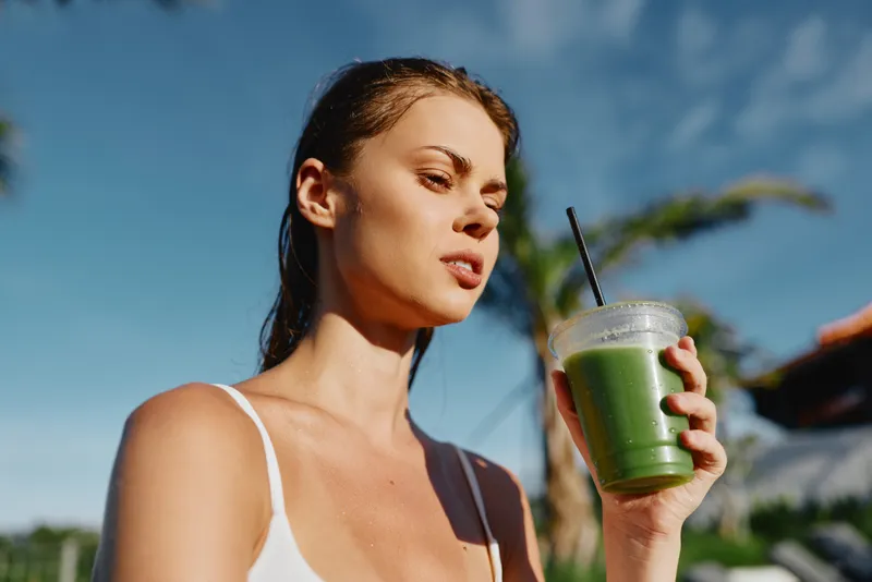 Smiling woman in a white tank top enjoys a green smoothie outdoors under a sunny sky. Wellness lifestyle and self care are highlighted by a bright, fresh mood.