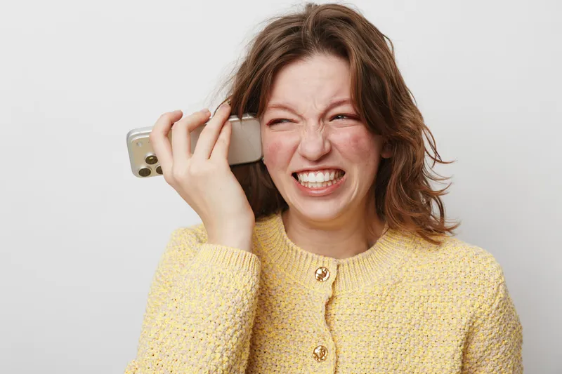 Frustrated young woman in yellow sweater holds smartphone near ear, grimacing with anger. Isolated on white background. Concept of bad news, communication problems, and stress.
