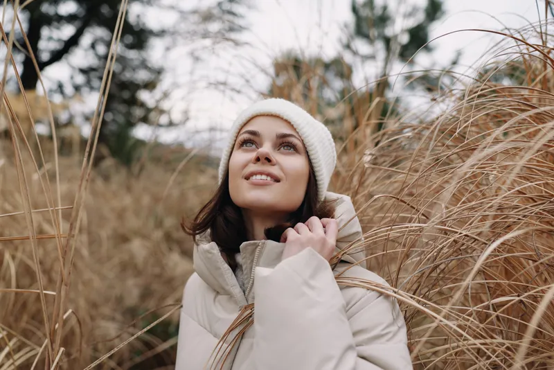 Woman in a warm coat and knit hat stands among tall grasses, gazing upward with a soft smile, in a natural field setting during a calm outdoor day.