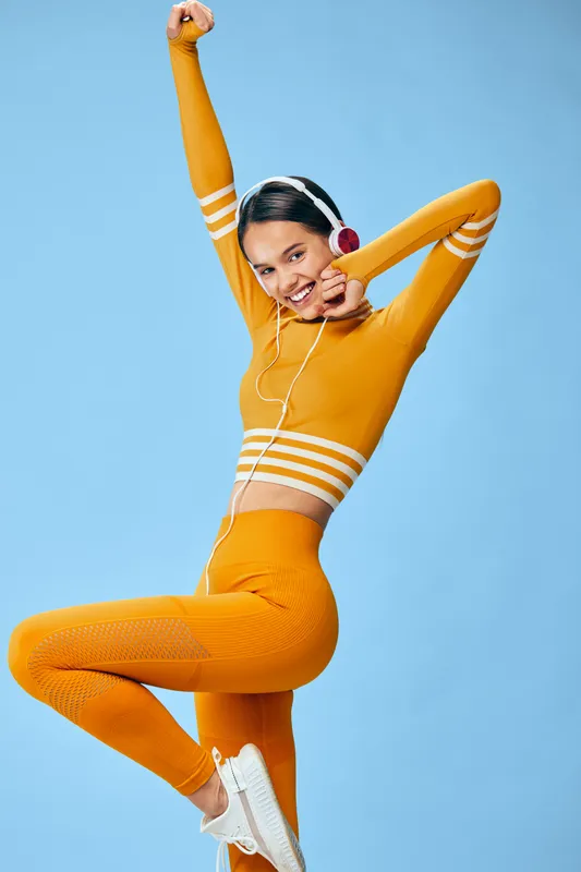 Young woman fitness model in a bright orange seamless workout set stretching with headphones and white sneakers, energetic sport pose against a clean blue studio background.