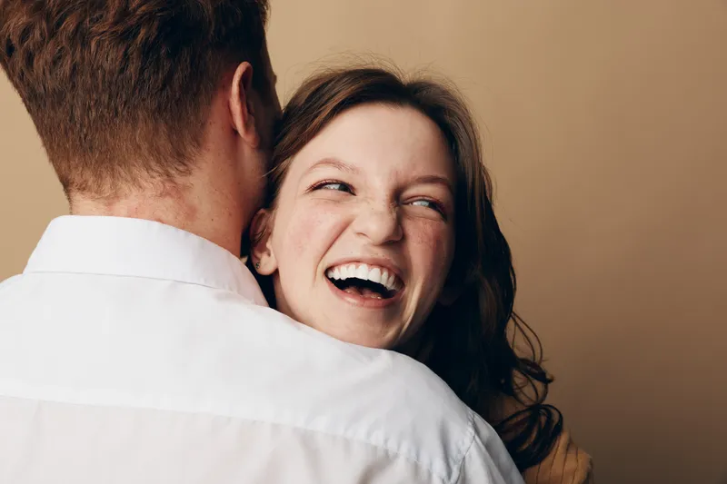 Happy young woman with curly hair laughing while hugging a man in white shirt. Close-up portrait on beige background. Concept of joy, affection, and relationship moments.