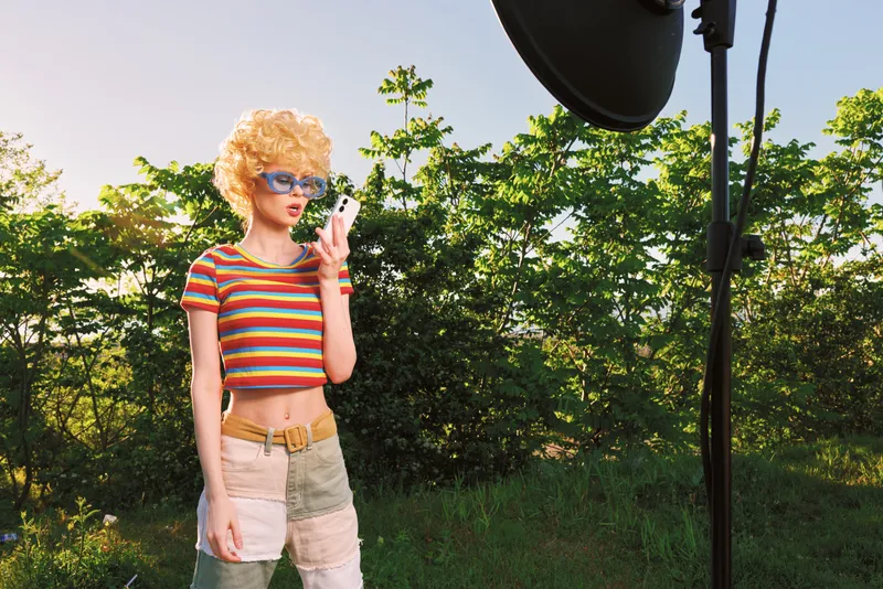 Smiling woman with curly blonde hair in a striped crop top and shorts. Outdoor studio lighting highlights vibrant retro film color, casual fashion, and confident lifestyle.
