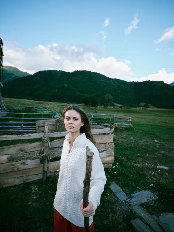Woman in a white knitted sweater stands beside a wooden fence in a wide countryside field, carrying a wooden stick, with green hills and blue sky behind