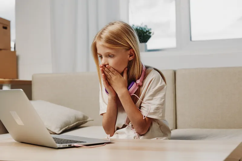 Surprised young girl with headphones covers her mouth looking at laptop screen in bright living room. Concept for online surprise, technology, childhood, and remote learning emotions.