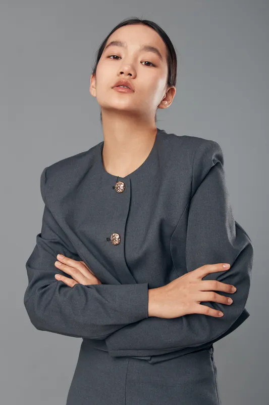 Young woman with folded arms in stylish gray blazer looking confidently at camera on plain background. Portrait, fashion, attitude, professional, female model with natural makeup and slicked back